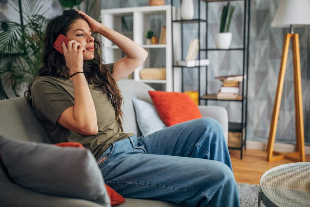 Woman sitting on a couch holding her head in stress while talking on a red phone in a cozy, modern living room with plants, shelves, and a lamp in the background.